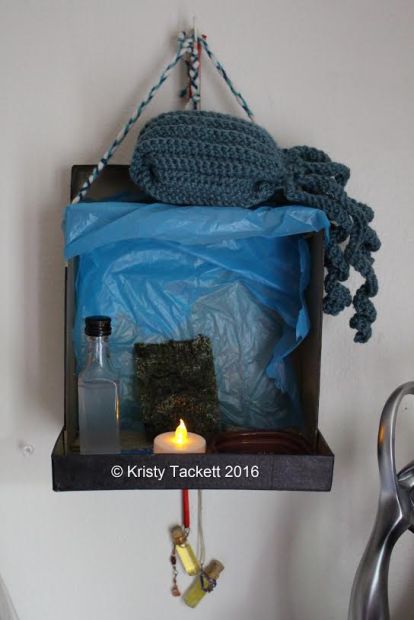 Hearth Keeper writes: This is a second shrine for Poseidon that I made from recycled materials that looks like a beach (more so on the inside). It's not quite finished, but then what shrine is in the beginning? On top is an octopus that I crocheted for Him. Within is a mini alcohol bottle filled with salt water, a piece of roasted seaweed, a red plate of rosemary and sea salt, an an LED light (because having a real tealight in this space isn't space). It's hanging by three braided yarns, each color representing Poseidon, Zeus, and Hades. Hanging down beneath are protective bottles. Not pictured in this photo is a dolphin charm that represents His wife, Amphitrite. [Jo writes: YAY POSEIDON SHRINE!!!! *ahem*]